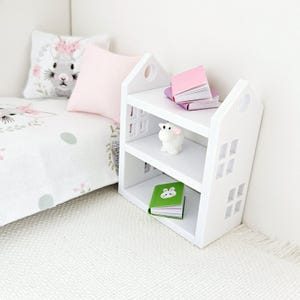 May include: White bookshelf with two shelves, featuring a small white lamb figurine, a green book with a bunny illustration, and two pink books stacked on top of each other. The bookshelf is in a dollhouse bedroom setting.