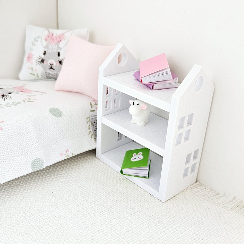 May include: White bookshelf with two shelves, featuring a small white lamb figurine, a green book with a bunny illustration, and two pink books stacked on top of each other. The bookshelf is in a dollhouse bedroom setting.