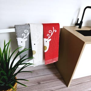 May include: Three kitchen towels hanging on a rack. The towels are gray, white, and red with a white reindeer design.
