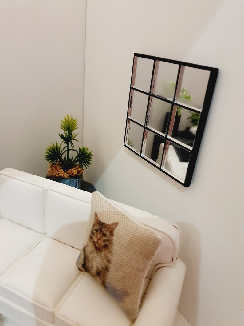 May include: A white couch with a pillow featuring a cat design sits in a room with a black framed mirror with nine square panes. A small potted plant is on the floor next to the couch.