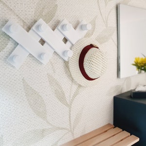 以下が含まれることがあります： 5つのフックが付いた白い壁掛けのハンガーラック、フックの1つに赤いバンドが付いた麦わら帽子がかけられており、小さな木のベンチと2つの引き出しが付いた濃い灰色のドレッサーがあります。