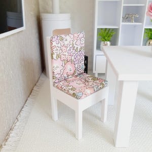 Puede incluir: Silla de madera blanca con un cojín de estampado floral. La silla tiene un diseño sencillo con cuatro patas y un asiento cuadrado.