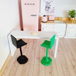 May include: A white table with two bar stools, one black and one green. The table has a plate of pink and yellow pastries on it. The table is in a dollhouse kitchen setting.