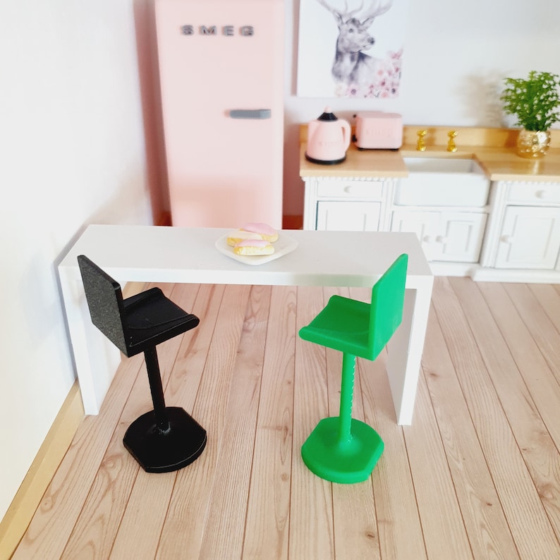 May include: A white table with two bar stools, one black and one green. The table has a plate of pink and yellow pastries on it. The table is in a dollhouse kitchen setting.