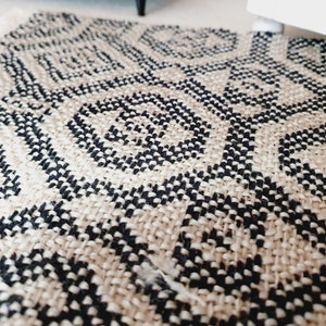 May include: Close-up of a woven rug with a geometric pattern in black and beige. The rug's design features repeating diamond and circular shapes. The rug has a fringed edge and is made of a natural fiber.