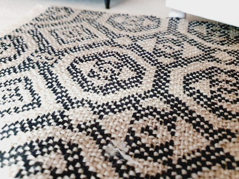 May include: Close-up of a woven rug with a geometric pattern in black and beige. The rug's design features repeating diamond and circular shapes. The rug has a fringed edge and is made of a natural fiber.