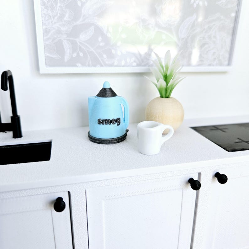 May include: A blue and black electric kettle with the word "Smeg" on the front, a white ceramic mug, and a small potted plant on a white countertop.