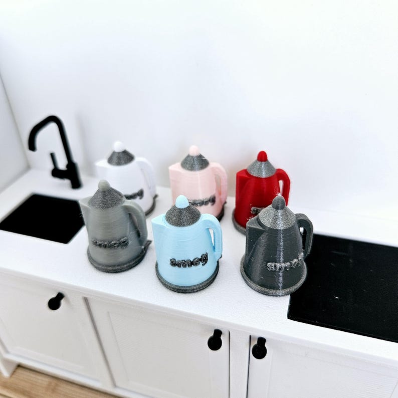 May include: Six miniature electric kettles in various colors, including white, pink, red, gray, and blue. The kettles are arranged in a row on a white countertop. Each kettle has the word "Smeg" printed on the side.