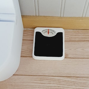 May include: A white bathroom scale with a black platform and a silver dial with red markings. The dial reads "0 120 10 2".