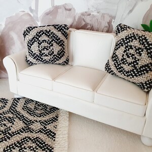 May include: Miniature white sofa with two decorative pillows. The pillows and rug feature a black and beige geometric pattern. The sofa is placed on a white rug, and a black cabinet is visible in the background. The scene evokes a modern, minimalist aesthetic.