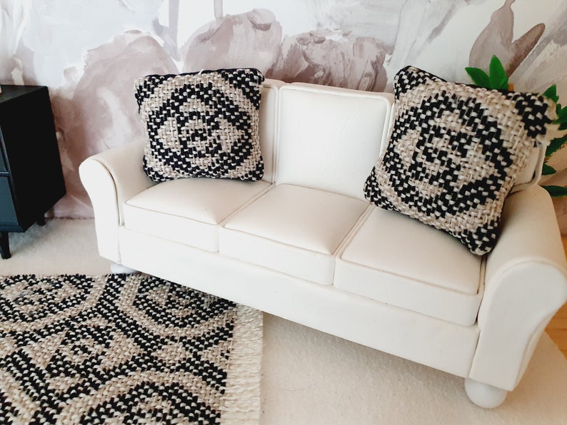 May include: Miniature white sofa with two decorative pillows. The pillows and rug feature a black and beige geometric pattern. The sofa is placed on a white rug, and a black cabinet is visible in the background. The scene evokes a modern, minimalist aesthetic.
