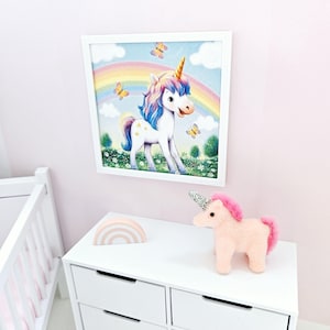 Pode incluir: Uma imagem emoldurada em branco de um unic&oacute;rnio com um arco-&iacute;ris atr&aacute;s. O unic&oacute;rnio est&aacute; em p&eacute; em um campo de flores. H&aacute; um unic&oacute;rnio de pel&uacute;cia rosa e branco em uma c&ocirc;moda branca com uma decora&ccedil;&atilde;o de madeira em forma de arco-&iacute;ris.