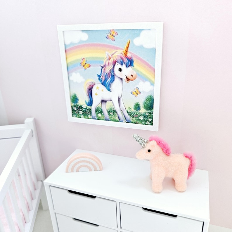 Pode incluir: Uma imagem emoldurada em branco de um unic&oacute;rnio com um arco-&iacute;ris atr&aacute;s. O unic&oacute;rnio est&aacute; em p&eacute; em um campo de flores. H&aacute; um unic&oacute;rnio de pel&uacute;cia rosa e branco em uma c&ocirc;moda branca com uma decora&ccedil;&atilde;o de madeira em forma de arco-&iacute;ris.