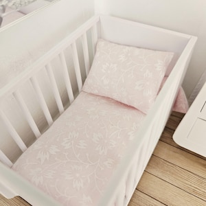 May include: A white crib with a light pink floral patterned pillow and fitted sheet. The bedding has a delicate white floral design. The crib is made of white wood and has vertical slats.