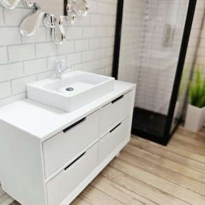 May include: A miniature white bathroom vanity with a rectangular sink and chrome faucet. The vanity has six drawers with black handles. A shower with a black frame is visible in the background. The floor is made of wood.