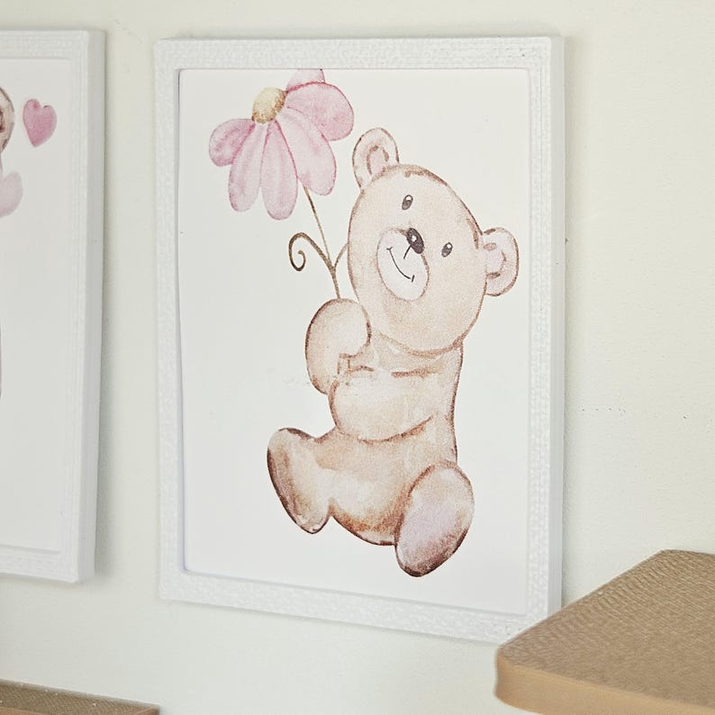 May include: A rectangular wall art piece in a white frame featuring a watercolour illustration of a light brown teddy bear holding a large pink flower with a yellow centre. The bear has a simple smiling face and is depicted in a seated position.