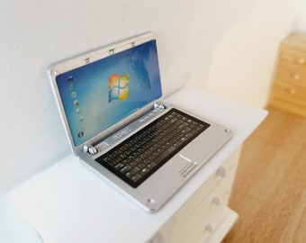 Puede incluir: Un ordenador portátil plateado con un teclado negro y una pantalla azul que muestra el logotipo de Windows. El portátil está sobre una superficie blanca.