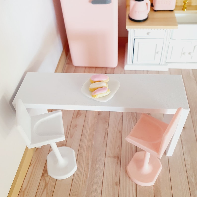 May include: A white table with two bar stools, one white and one pink. There are three pink and white eclairs on a white plate on the table.
