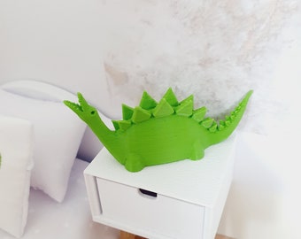May include: A bright green 3D printed dinosaur figurine with a spiky back and a long tail. The dinosaur is sitting on a white nightstand with a drawer.