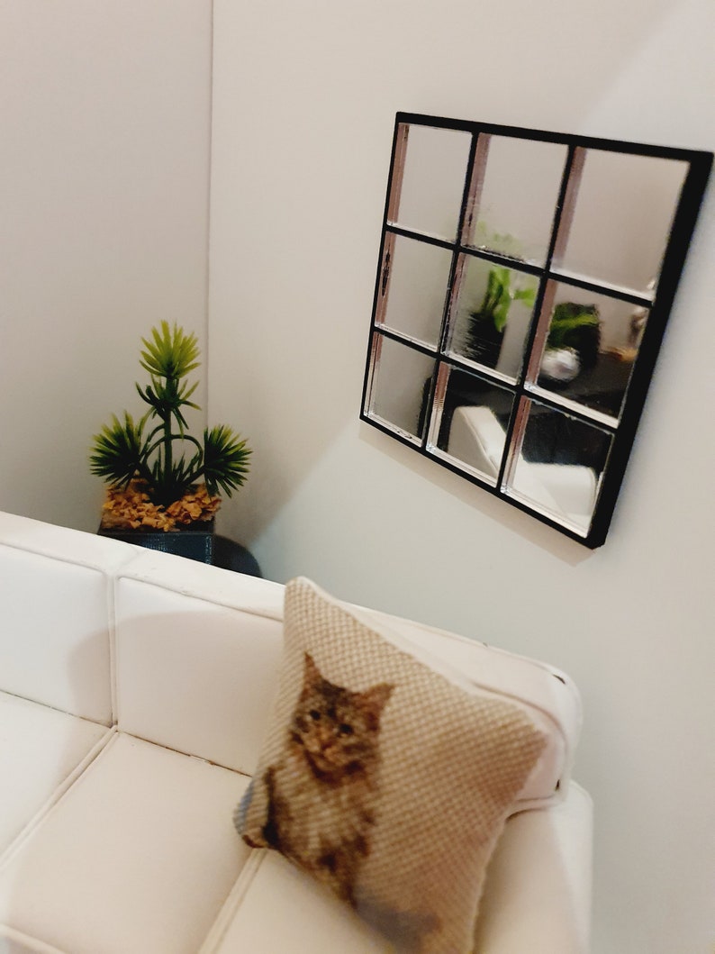 May include: A white couch with a pillow featuring a brown and white cat design. A small potted plant sits on a table in the background. A black framed mirror with multiple panes hangs on the wall.