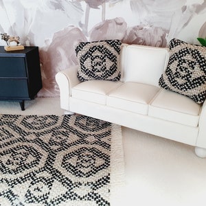 May include: Miniature living room set with a white sofa, two patterned pillows, and a black and white rug. A black cabinet and decorative items complete the scene. The rug has a geometric design.