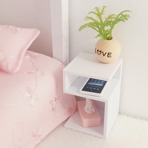 May include: White bedside table with two shelves. The top shelf has a small potted plant with the word "LOVE" on the pot. The bottom shelf has a pink tissue box and a black tablet.
