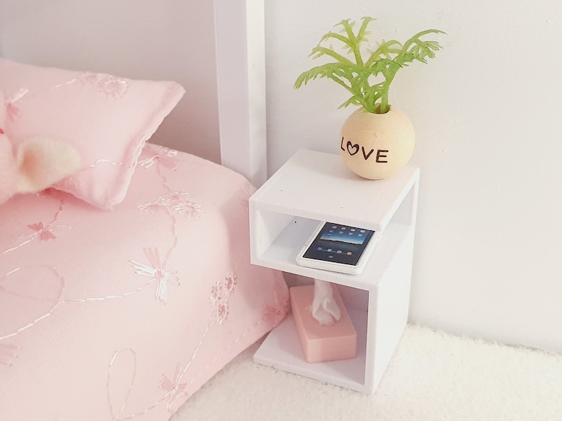 May include: White bedside table with two shelves. The top shelf has a small potted plant with the word "LOVE" on the pot. The bottom shelf has a pink tissue box and a black tablet.