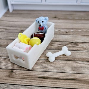 May include: A white toy storage bin with cut-out paw prints on the sides. The bin is filled with toys, including a teddy bear, a football, a yellow ball, and a bone.