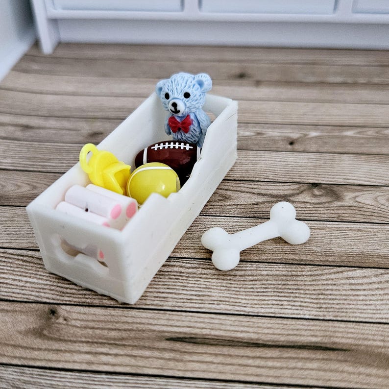 May include: A white toy storage bin with cut-out paw prints on the sides. The bin is filled with toys, including a teddy bear, a football, a yellow ball, and a bone.
