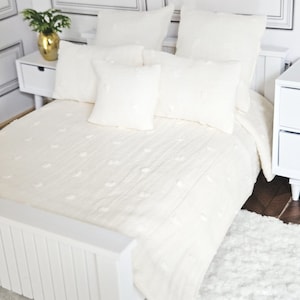 May include: A cream-coloured bed with a matching duvet and pillows. The duvet and pillows have a textured, dotted pattern. The bed is flanked by white bedside tables and a fluffy white rug. A gold vase with greenery sits on one bedside table.