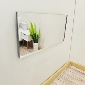 May include: A rectangular, frameless mirror mounted on a white wall. The mirror reflects a white nightstand with a drawer, a green plant in a white vase, and a portion of a white wall. The corner of the room has light wood flooring.