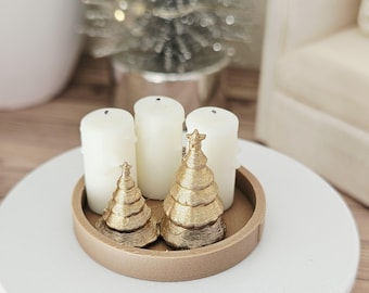 May include: A round tan tray containing three white pillar candles and two gold-colored, textured, cone-shaped tree figurines. The smaller tree is positioned to the left, while the larger tree, topped with a star, sits to the right.