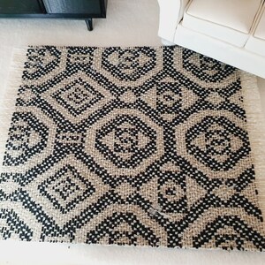 May include: A small, rectangular rug with a geometric diamond pattern in black and beige. The rug has a fringed edge and is placed on a white surface. The rug is near a white sofa and a black cabinet.