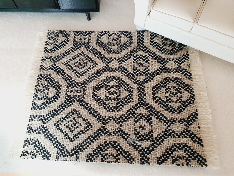 May include: A small, rectangular rug with a geometric diamond pattern in black and beige. The rug has a fringed edge and is placed on a white surface. The rug is near a white sofa and a black cabinet.