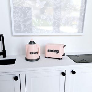 May include: A pink miniature kettle and toaster with the word "smeg" printed on them. The appliances are on a white countertop with black cabinet doors.