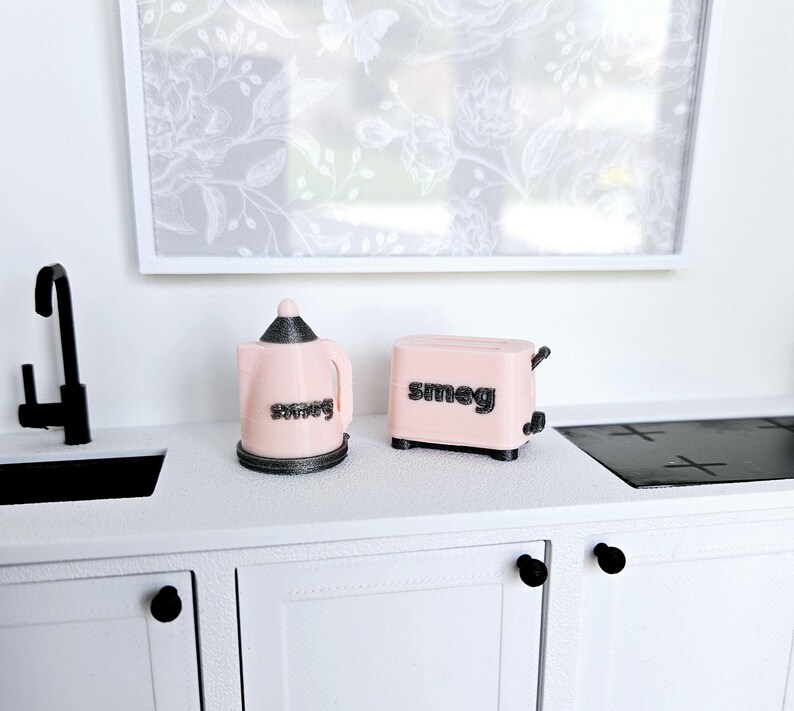 May include: A pink miniature kettle and toaster with the word "smeg" printed on them. The appliances are on a white countertop with black cabinet doors.
