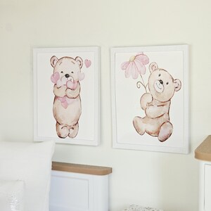 May include: Two rectangular wall art prints in white frames featuring watercolour illustrations of light brown teddy bears. One bear holds a cluster of pink hearts, while the other bear holds a single large pink flower against a white background.