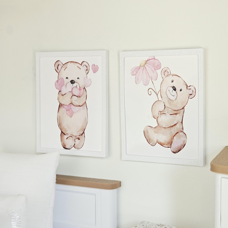 May include: Two rectangular wall art prints in white frames featuring watercolour illustrations of light brown teddy bears. One bear holds a cluster of pink hearts, while the other bear holds a single large pink flower against a white background.