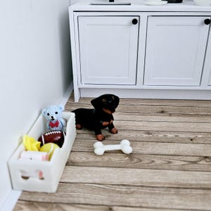 May include: A white toy storage bin with a blue teddy bear, a red and white football, and a yellow and white toy inside. A black and brown dachshund puppy figurine is standing next to the bin with a white bone toy in front of it.