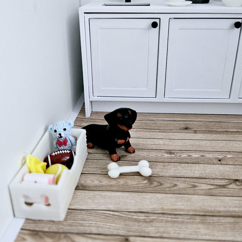 May include: A white toy storage bin with a blue teddy bear, a red and white football, and a yellow and white toy inside. A black and brown dachshund puppy figurine is standing next to the bin with a white bone toy in front of it.