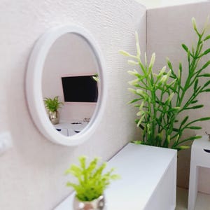 May include: A white round mirror with a white frame hangs on a white wall. The mirror reflects a room with a black television, a potted fern, and a white dresser. A small potted plant sits on a white table in the foreground.