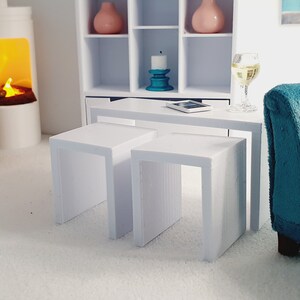 May include: White nesting tables with a white top and a teal blue candle holder on a white shelf in the background. A glass of white wine is on the table next to a teal blue sofa.