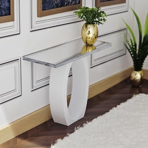 Op de afbeelding: Een moderne witte console met een helder glazen blad. De tafel heeft een uniek, gebogen basisontwerp. Een gouden vaas met een groene plant staat op de tafel. De tafel staat tegen een witte muur met ingelijste kunstwerken.