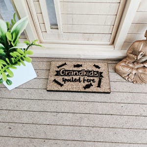 Puede incluir: Un letrero rectangular de corcho con el texto negro "Grandkids spoiled here", decorado con pequeñas formas negras. El letrero está sobre una superficie marrón claro, cerca de una pequeña planta y una estatua.