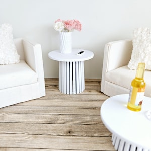 Op de afbeelding: Witte bijzettafel met een geflute basis en een witte vaas met roze en witte bloemen erop. De tafel staat in een woonkamer met twee witte banken en een houten vloer.