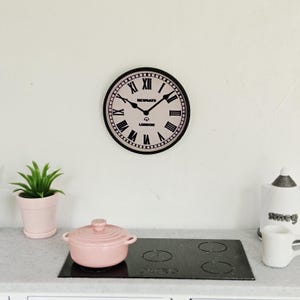 Puede incluir: Una escena de cocina en miniatura con un reloj rosa que muestra el texto "NEWGATE LONDON", una olla rosa, una placa de cocina negra y una pequeña planta en maceta. El reloj tiene números romanos y manecillas negras.