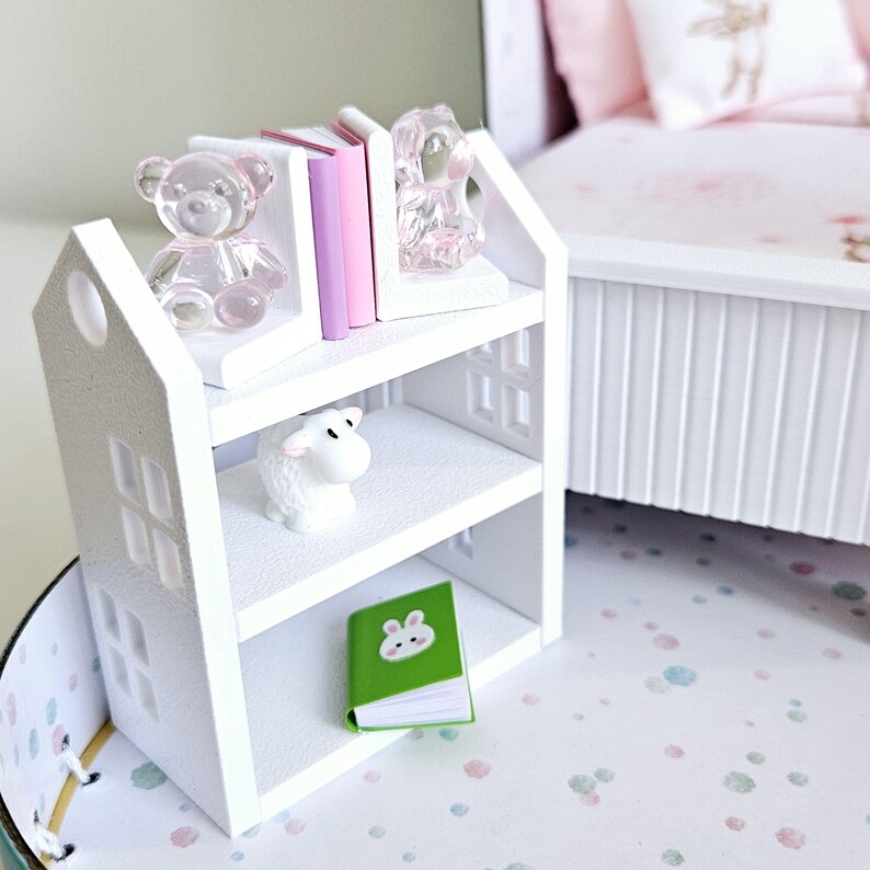 May include: White wooden bookshelf with three shelves. The top shelf has two pink and white bookends and a clear glass bear figurine. The middle shelf has a white sheep figurine. The bottom shelf has a green book with a bunny illustration.