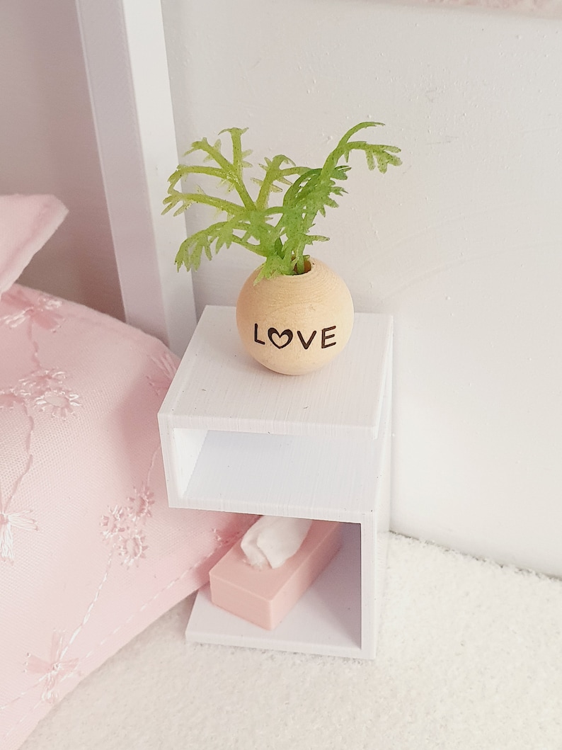May include: White bedside table with two shelves. The top shelf has a small wooden planter with the word "LOVE" inscribed on it. The planter has a green plant inside. The bottom shelf has a pink tissue box.