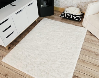 May include: A rectangular, off-white rug with a shaggy texture. The rug is placed on a wooden floor, and is surrounded by miniature furniture, including a white cabinet, a black fireplace, and a white armchair.