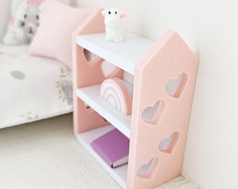 May include: A pink wooden shelf with heart cutouts and white shelves. The shelf is decorated with a rainbow, a small white figurine, and a purple book.
